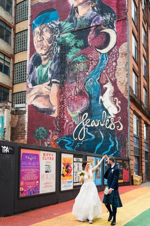 Wedding couple poses in front of vibrant street art mural on brick wall with colorful posters displayed below.