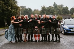 A wedding party poses together on a rainy day wearing traditional Scottish kilts and formalwear on a gravel path.