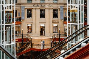 Interior of Cafe Bar 1780 shopping mall with escalators, ornate gold railings, and shoppers on multiple levels.