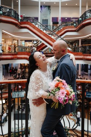Bride and groom embrace laughing in a multi-level shopping centre, bride holding a vibrant pink bouquet.