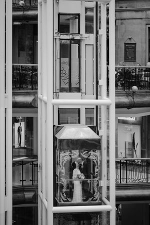 Black and white photo of a couple embracing in a glass elevator shaft of a modern building.
