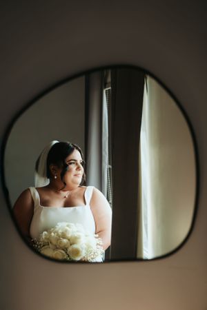 Artistic reflection in a curvy mirror shows a person in white holding a cream-colored bouquet while gazing out a window.