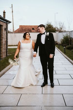 A couple stands together on a stone paved path at their wedding, she in a white mermaid dress and he in a black tuxedo.