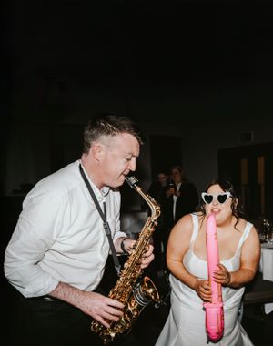 Two musicians perform at a party with one playing saxophone and another playing a pink recorder against a dark background.