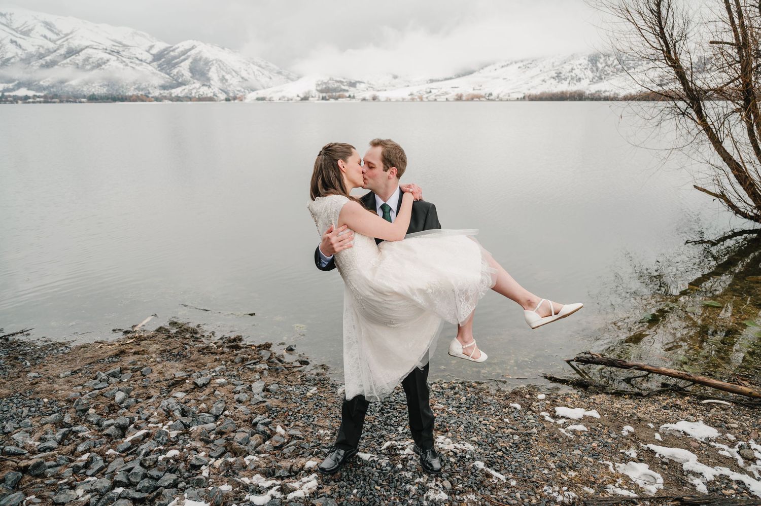 A couple embraces in a romantic pose on a snowy lakeshore with snow-capped mountains in the background.