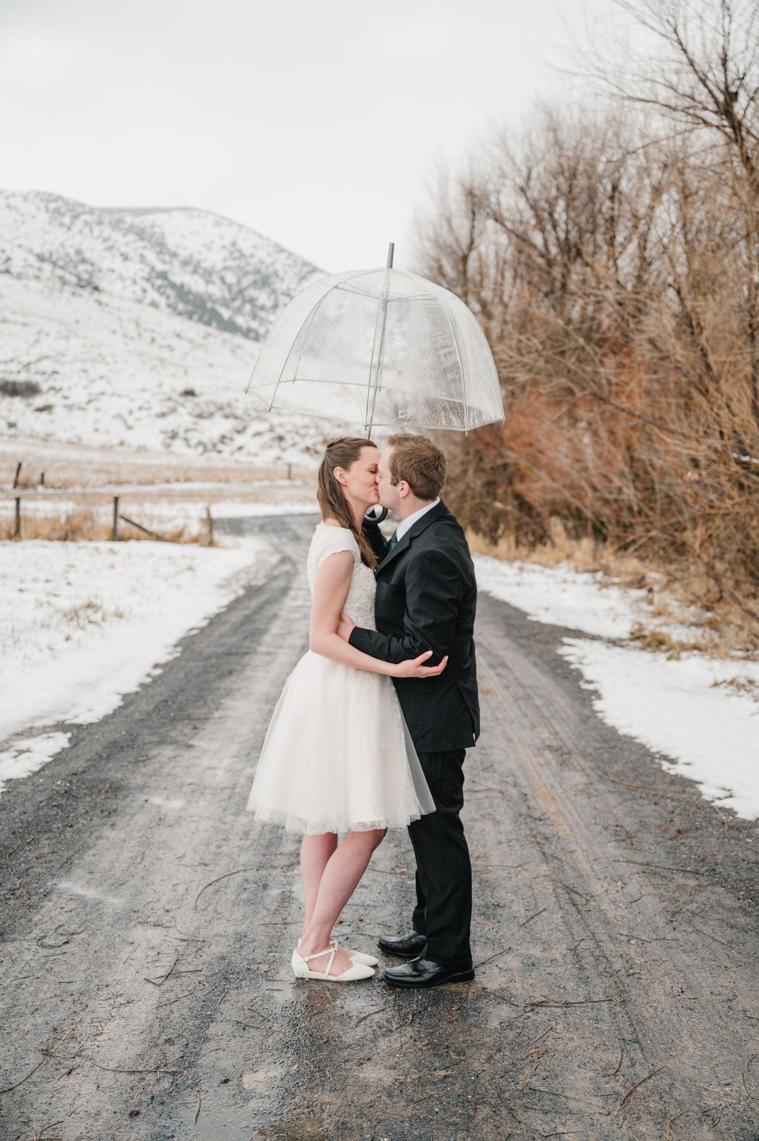A couple shares a romantic moment under a clear umbrella on a snowy mountain road during a winter wedding shoot.