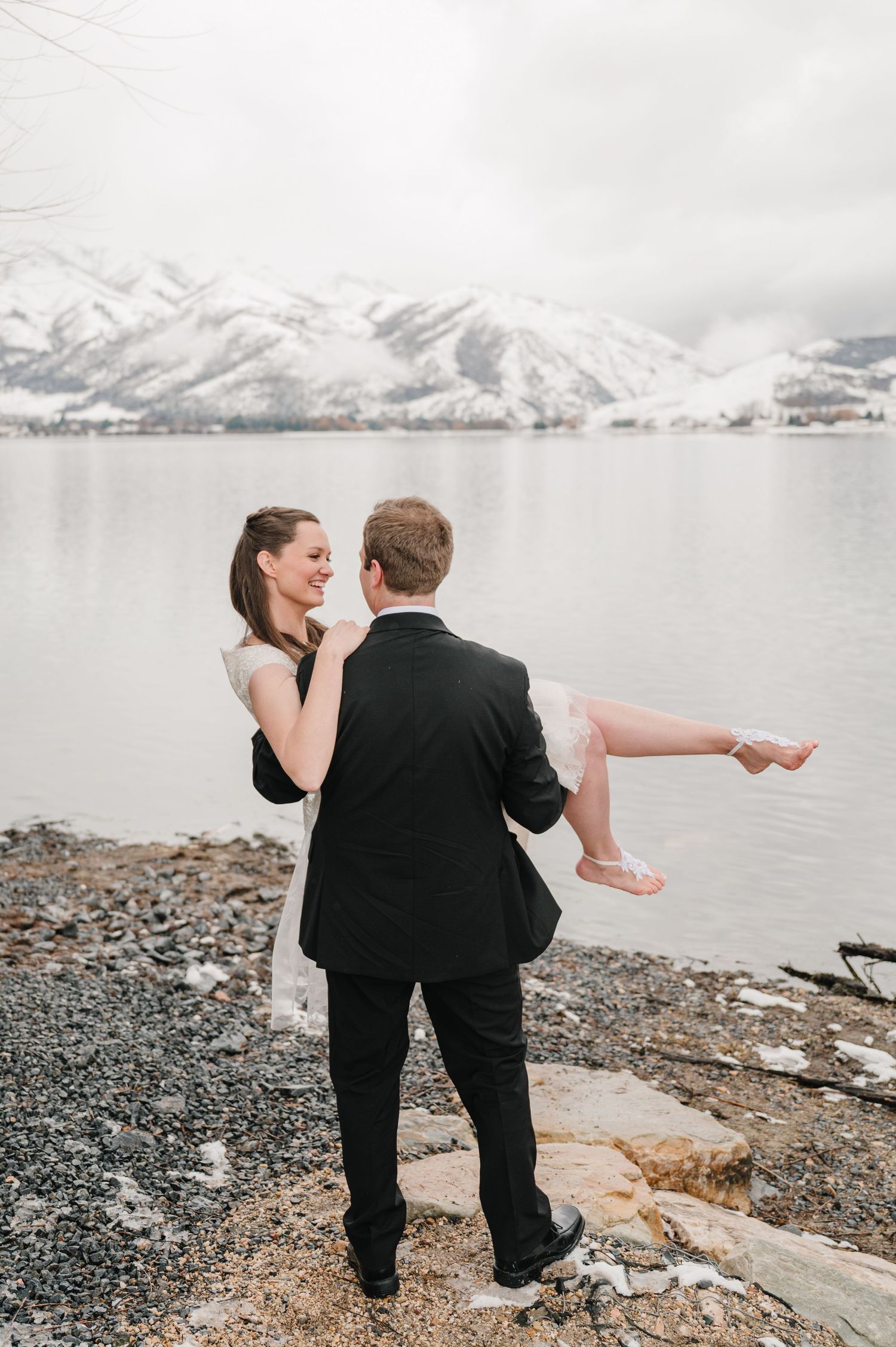 A couple shares a romantic moment by a snow-capped mountain lake as the groom carries his bride in formal attire.