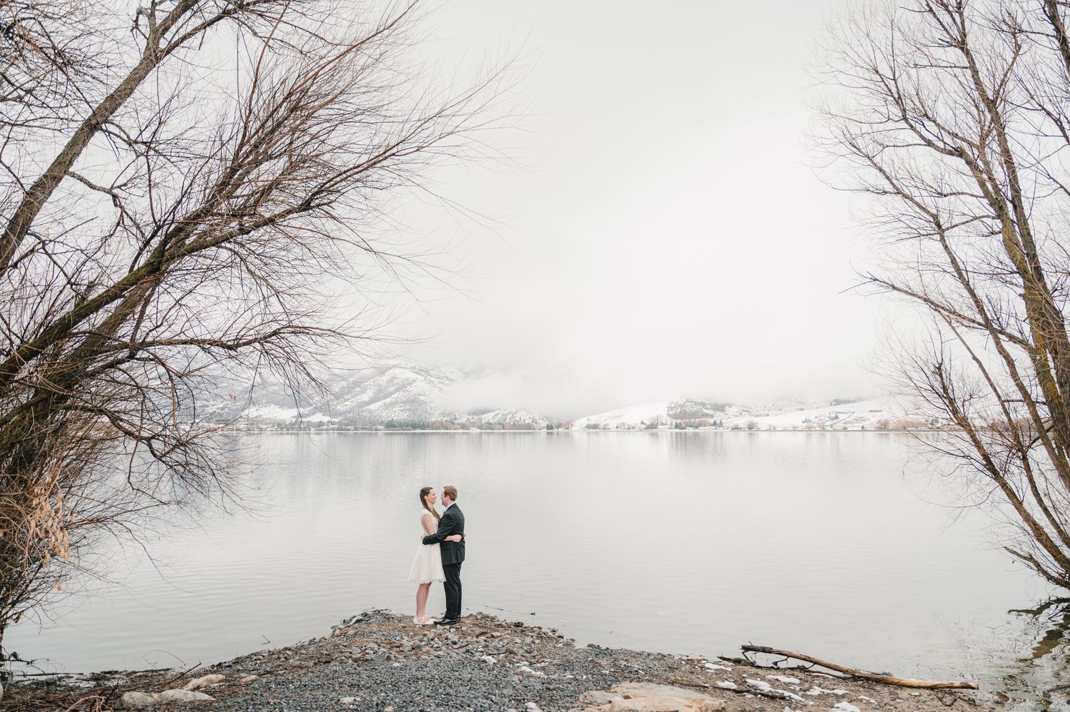 Two silhouettes embrace on a rocky outcrop overlooking a misty winter lake framed by bare trees.