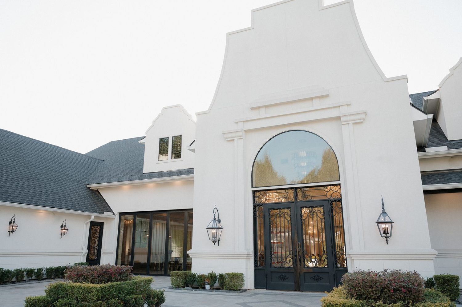 A white Spanish-style building entrance with grand wooden double doors, arched window, and elegant lantern sconces.