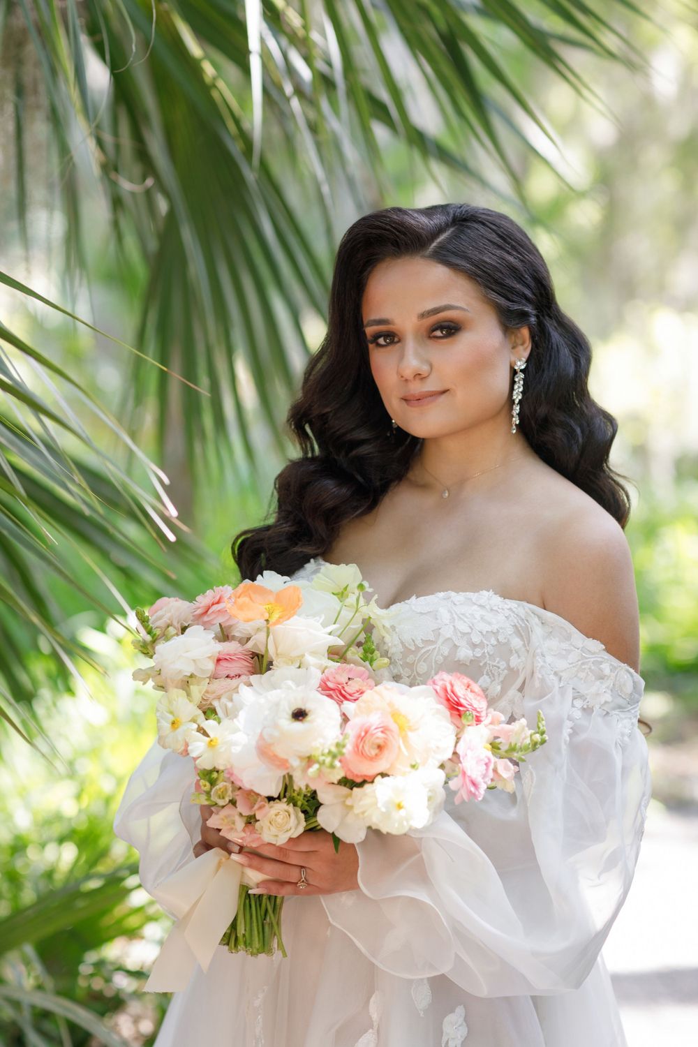 A bride in a white strapless gown with flowing sleeves holds a romantic bouquet of pink and peach flowers against palm leaves.