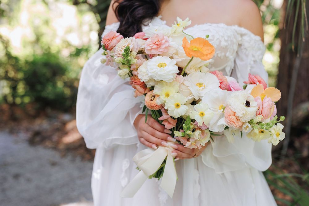 A delicate bouquet of pink, peach and white flowers with greenery held against a white wedding dress.