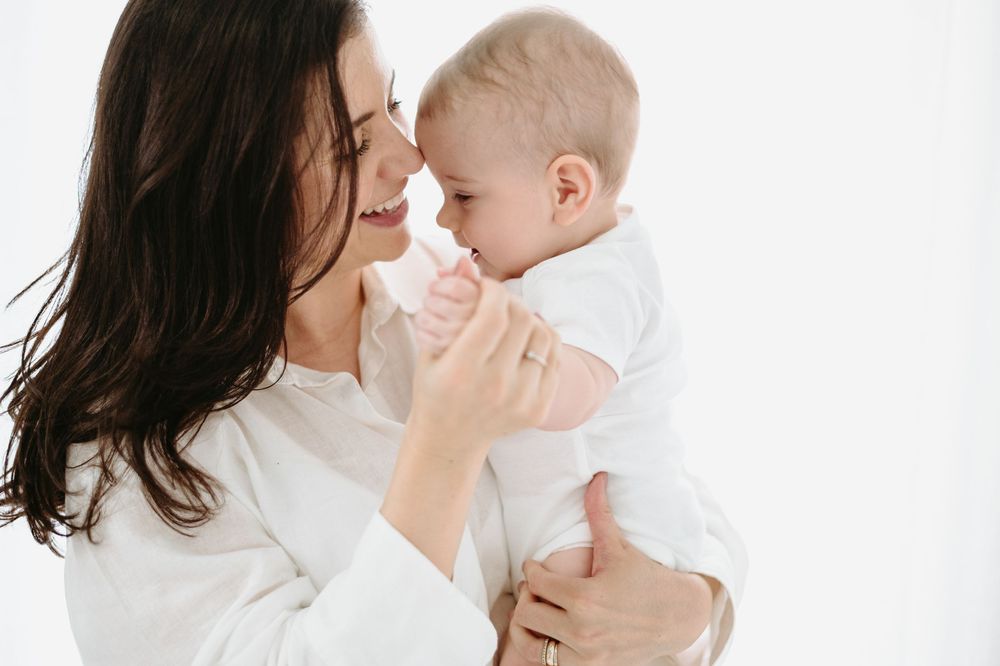 A joyful mother in white holds her smiling baby close, sharing a tender moment against a bright background.