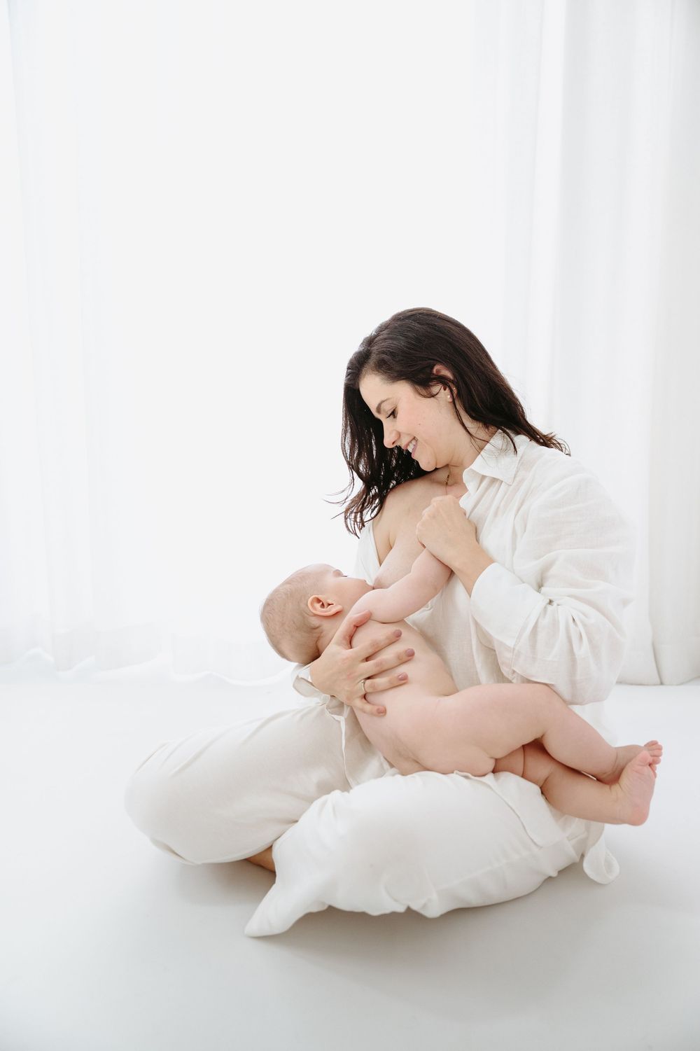 A mother breastfeeding her newborn baby while sitting on white pillows in a bright, serene white room.