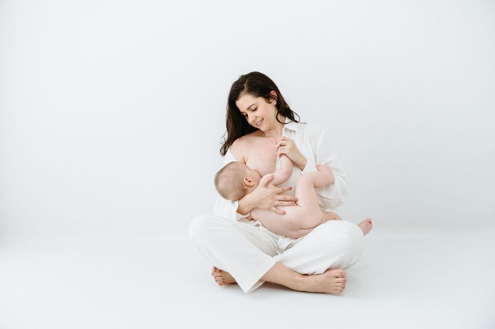 Mother in white outfit sitting cross-legged on white background, lovingly breastfeeding her newborn baby.