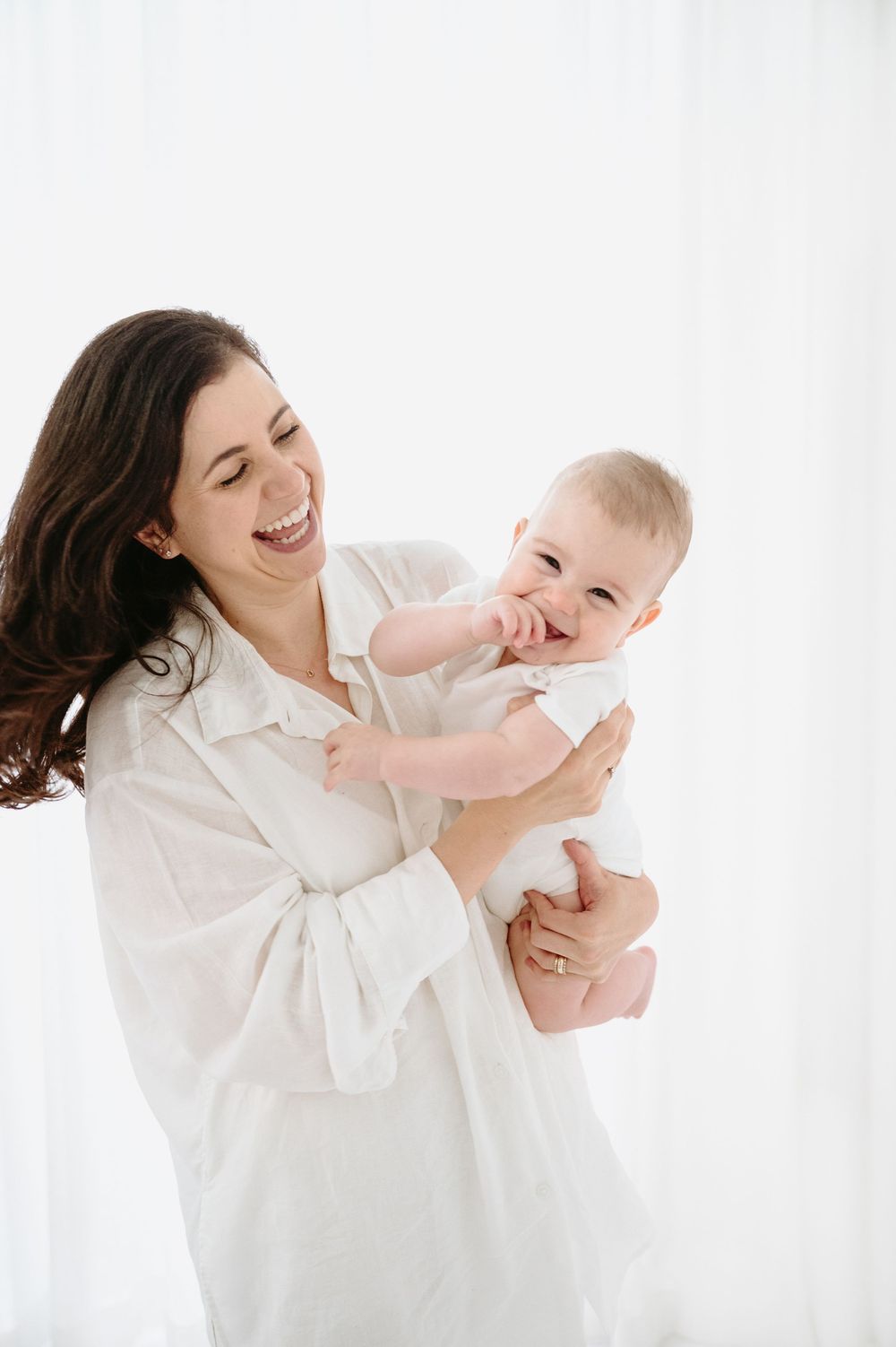 Happy mother in white shirt laughing while holding her smiling baby in a bright, airy white room.