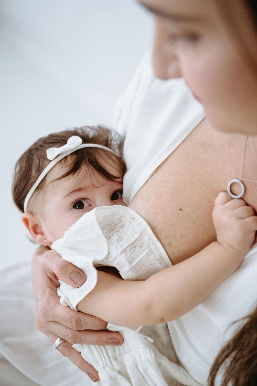 A mother in white dress breastfeeding her baby girl wearing a white bow headband and dress.