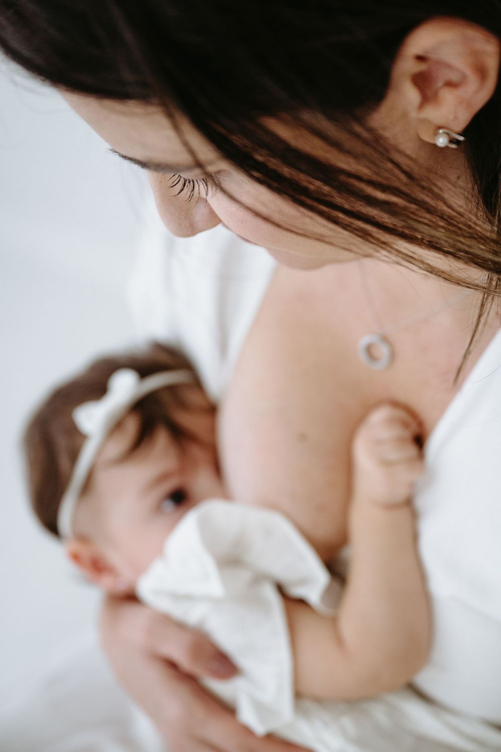 Mother in white dress breastfeeding her baby girl wearing a white bow headband.
