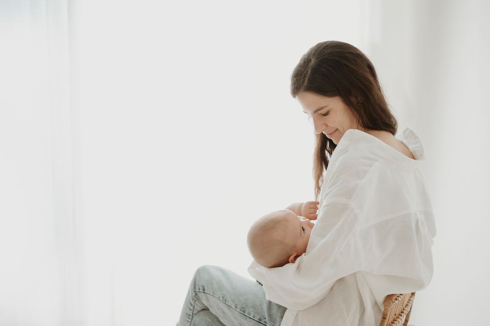 A mother breastfeeding her newborn baby while sitting in a chair near a bright window.