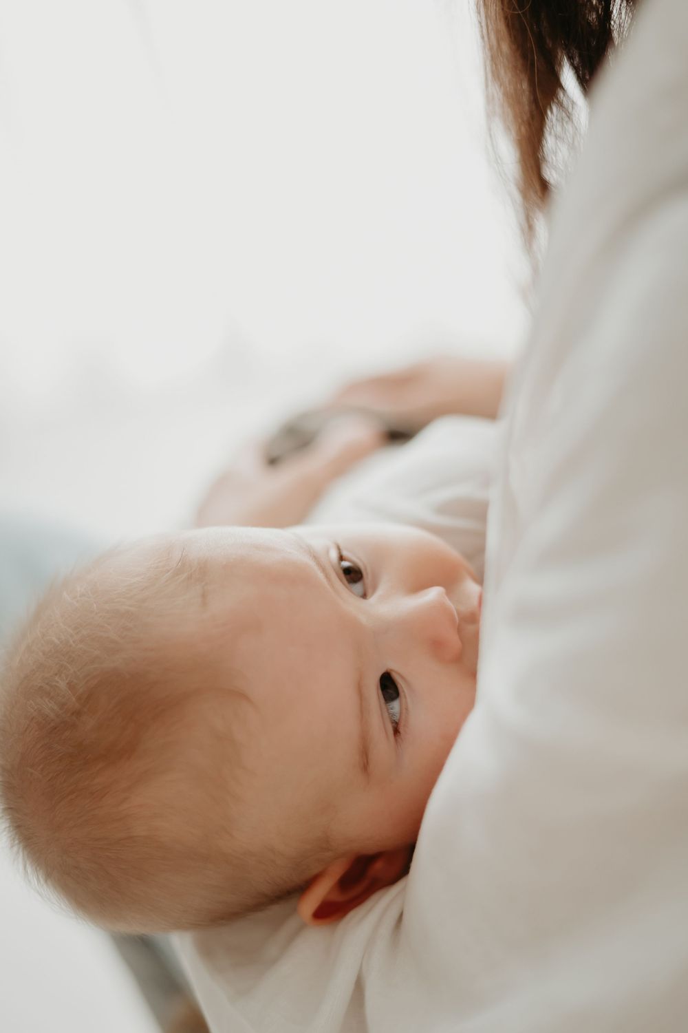 A newborn baby with blue eyes being cradled in a mother's arms, wearing a white onesie in soft natural light.