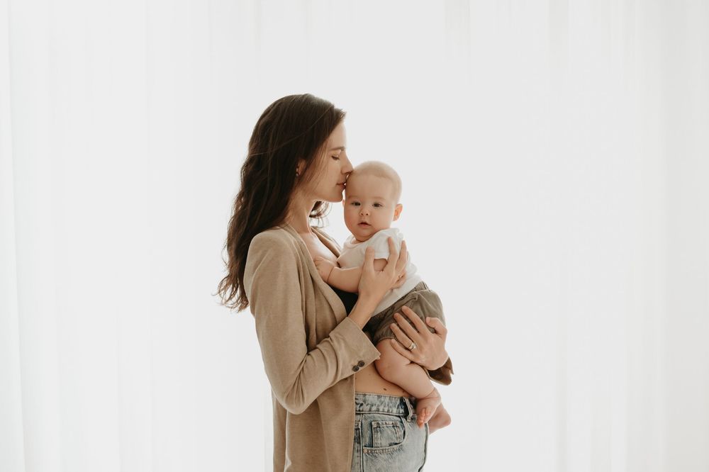 A mother tenderly kisses her baby while holding him against a soft white background.