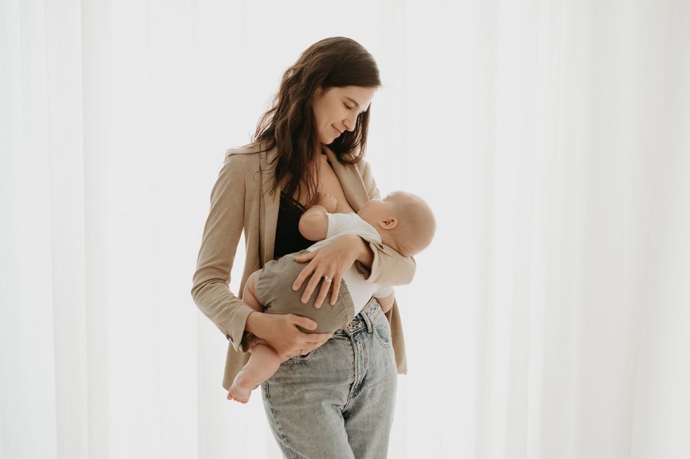 Young mother in beige blazer and jeans breastfeeding her newborn baby against a white curtain backdrop.
