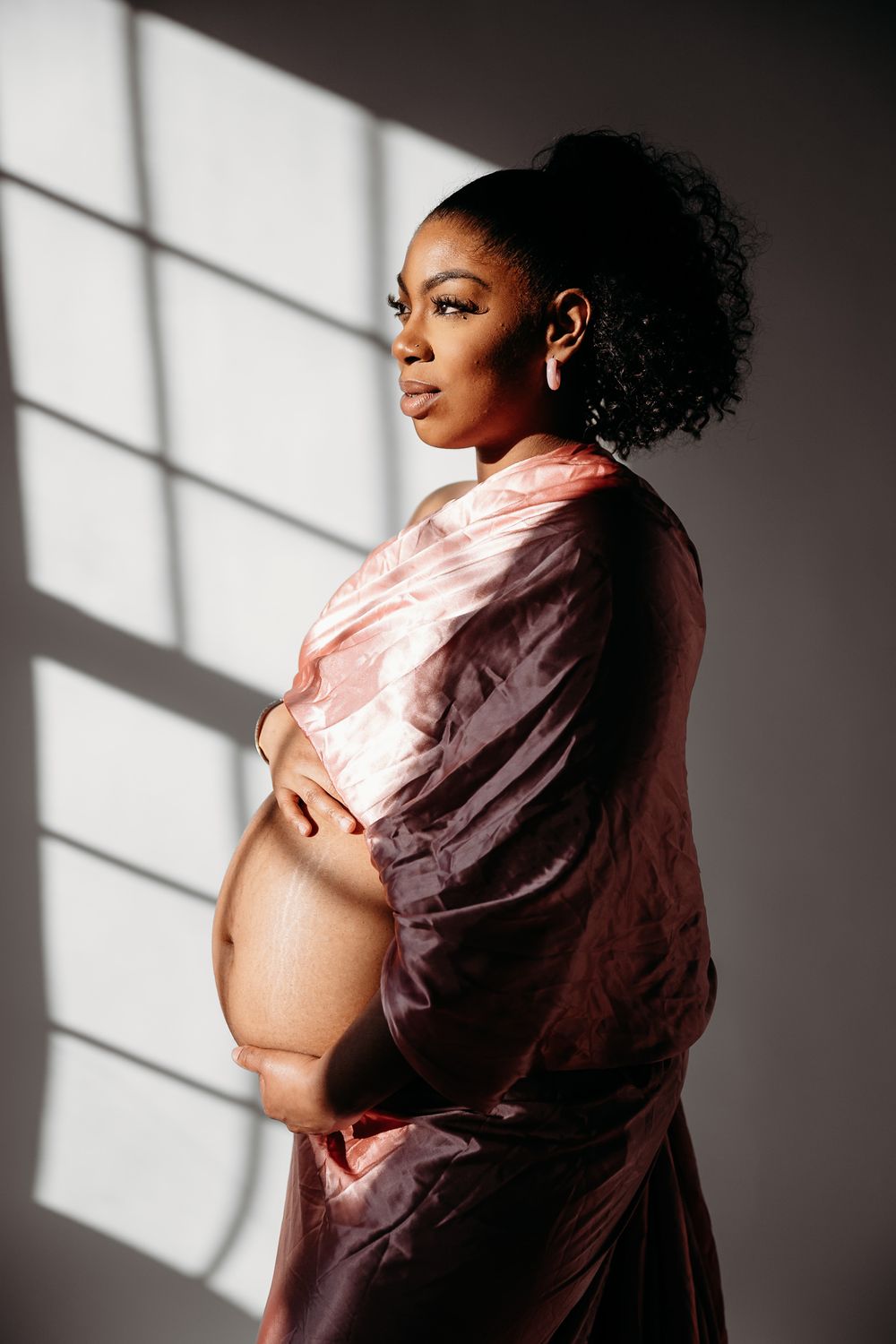 Artistic portrait with dramatic window light casting shadows on figure in pink flowing fabric.