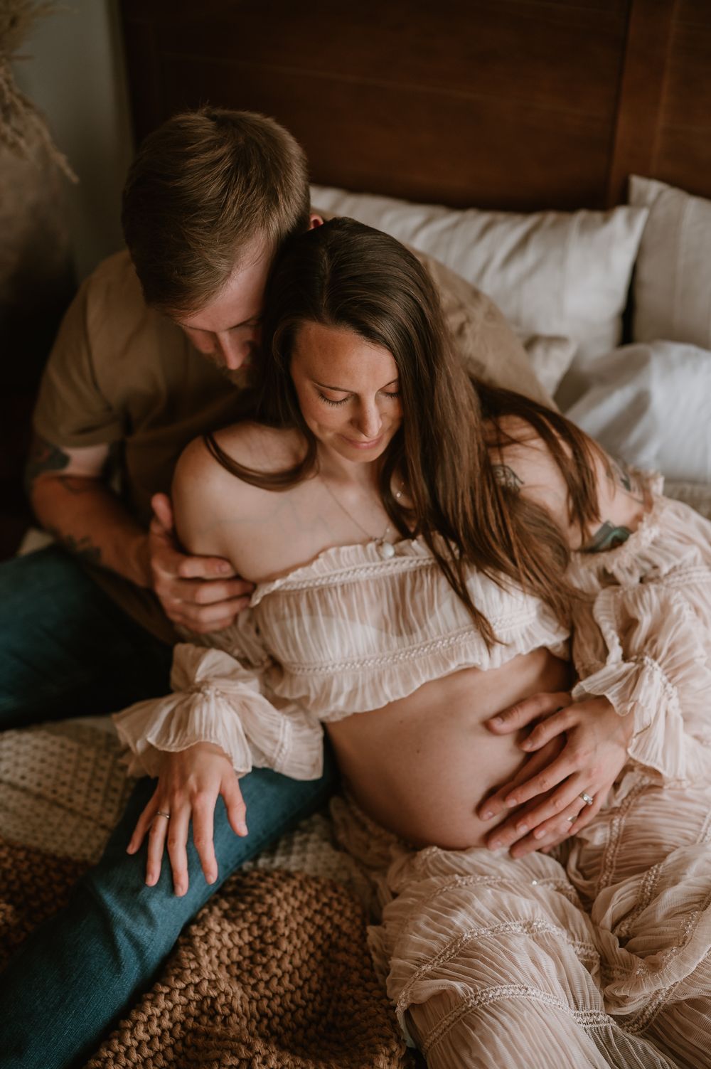 Intimate maternity photo showing a tender embrace on a bed with soft white linens and knit blanket.