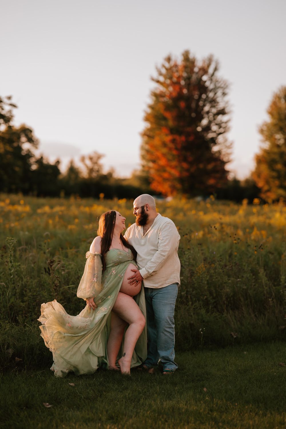 A romantic couple embraces in a golden autumn field at sunset with colorful fall foliage in the background.