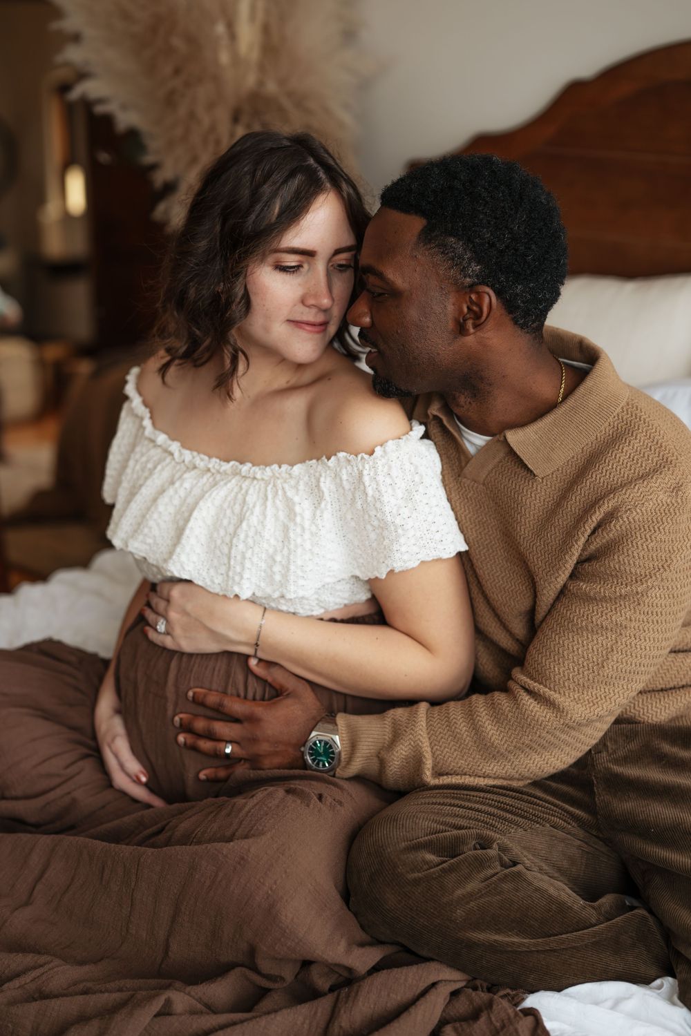 Intimate maternity moment shared on bed with brown bedding and wooden headboard decor.