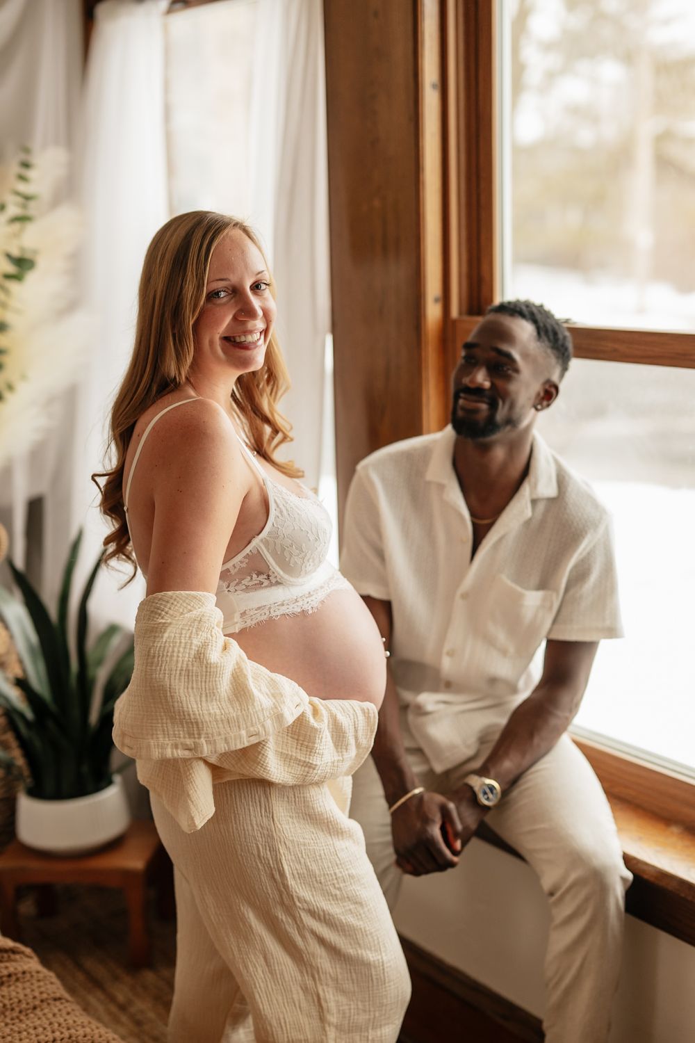 Expectant couple in white outfits share a tender moment by a sunny window with houseplants.