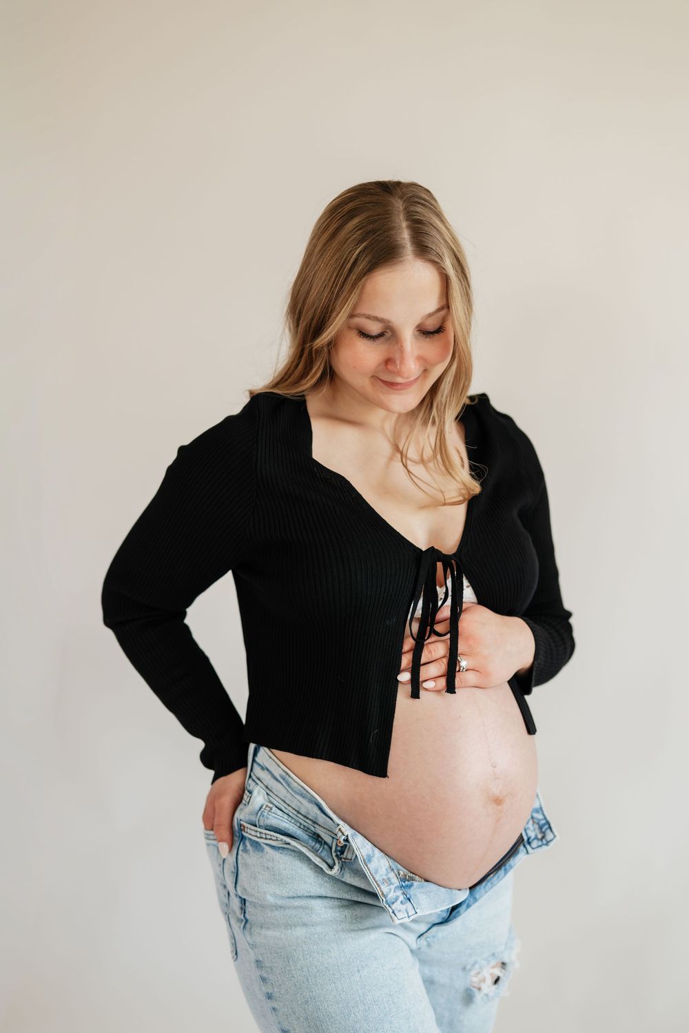 Smiling pregnant blonde woman in black crop top lifts shirt to reveal baby bump while wearing jeans.