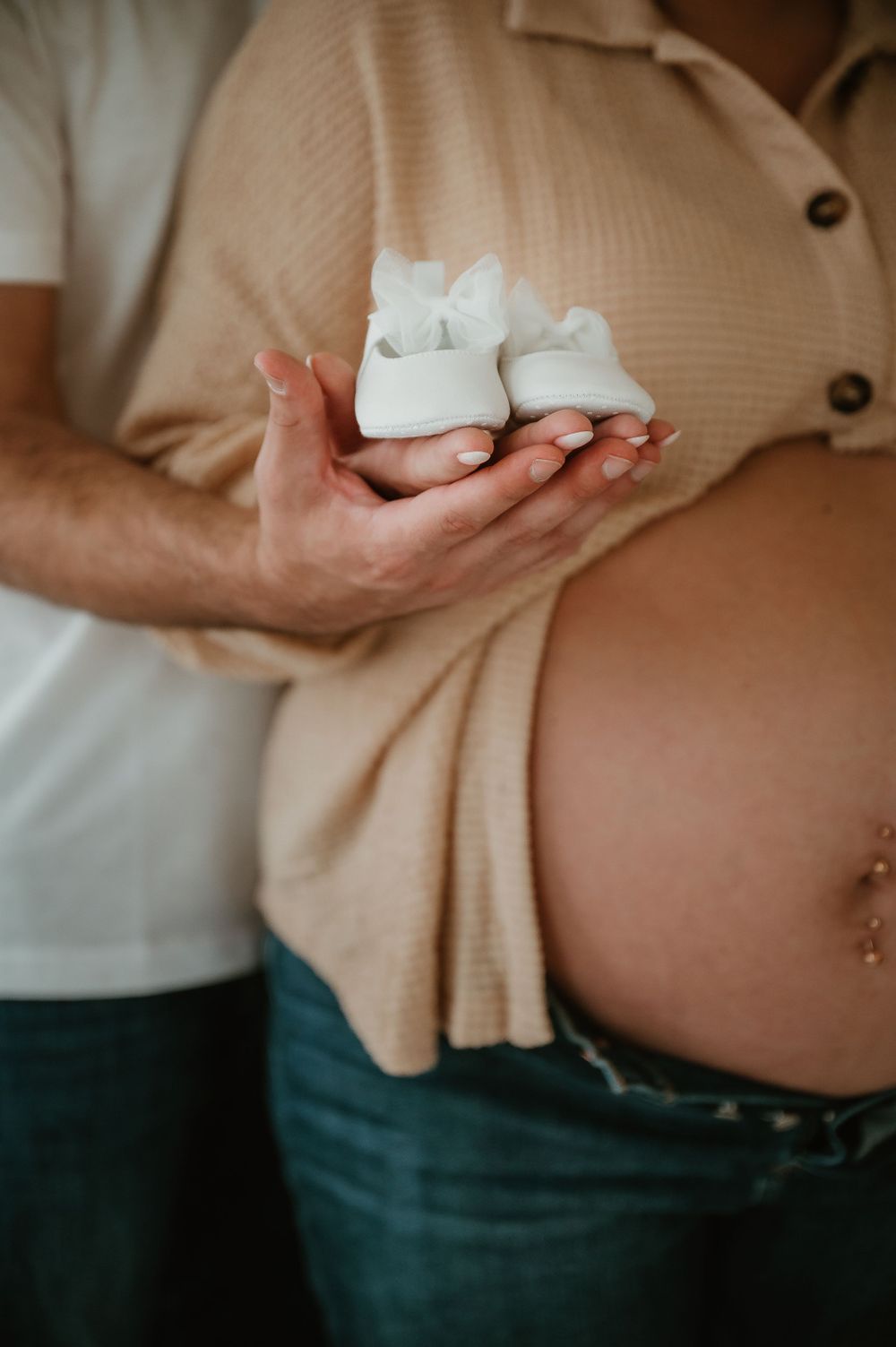 Hands holding tiny white baby shoes against a pregnant belly in a beige sweater.