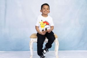 Child sitting on white ornate chair wearing Pokemon shirt against light blue background.