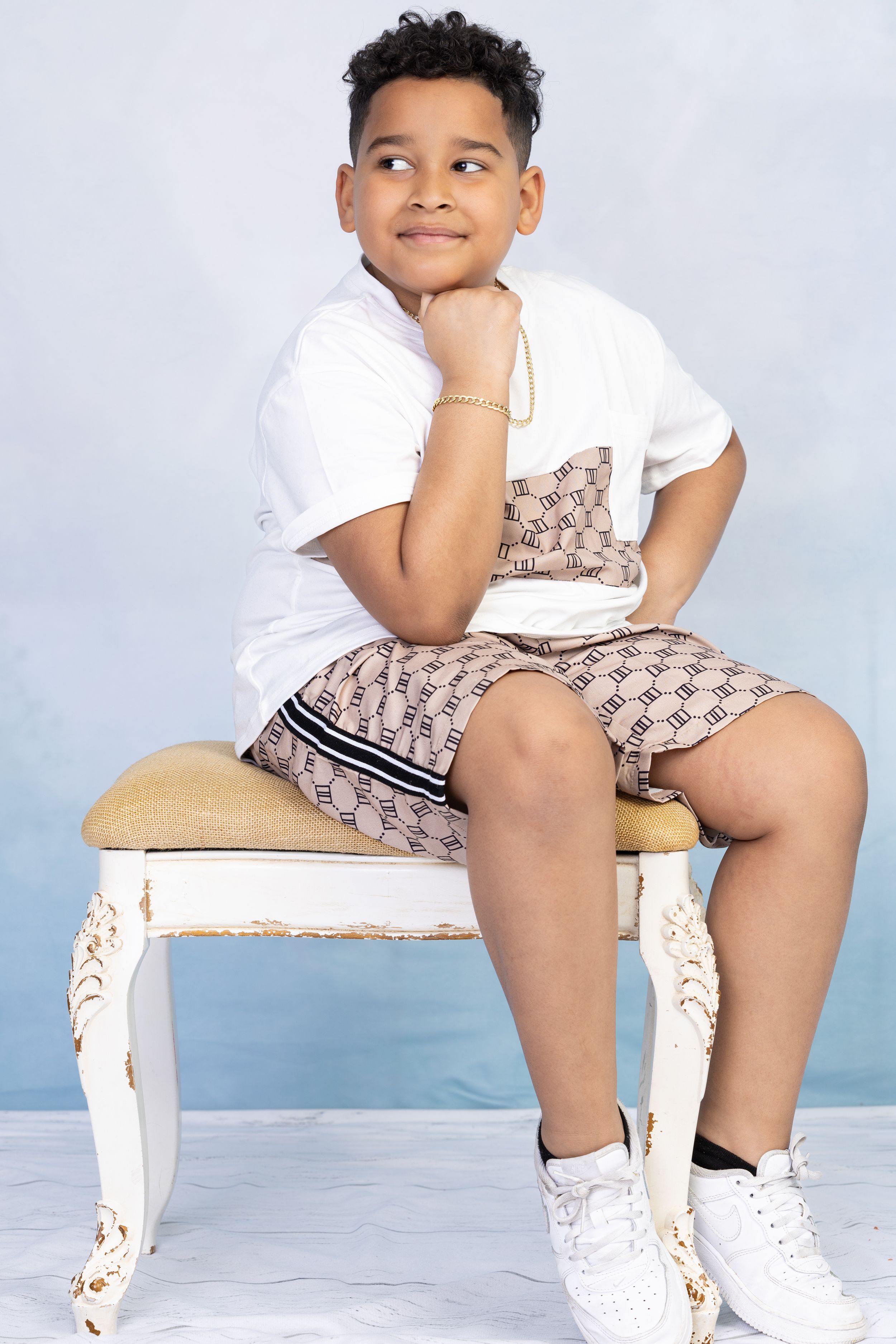 Young person wearing white t-shirt and patterned shorts sitting on ornate white chair.
