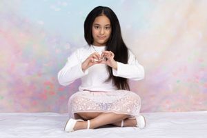 Long-haired figure in white top sitting cross-legged on floor with pastel background making heart shape with hands.
