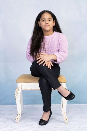 Child in pink sweater sitting on ornate white chair against blue backdrop.
