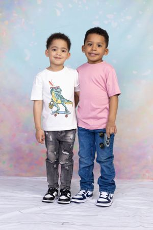 Two children wearing casual attire pose together against pastel background.