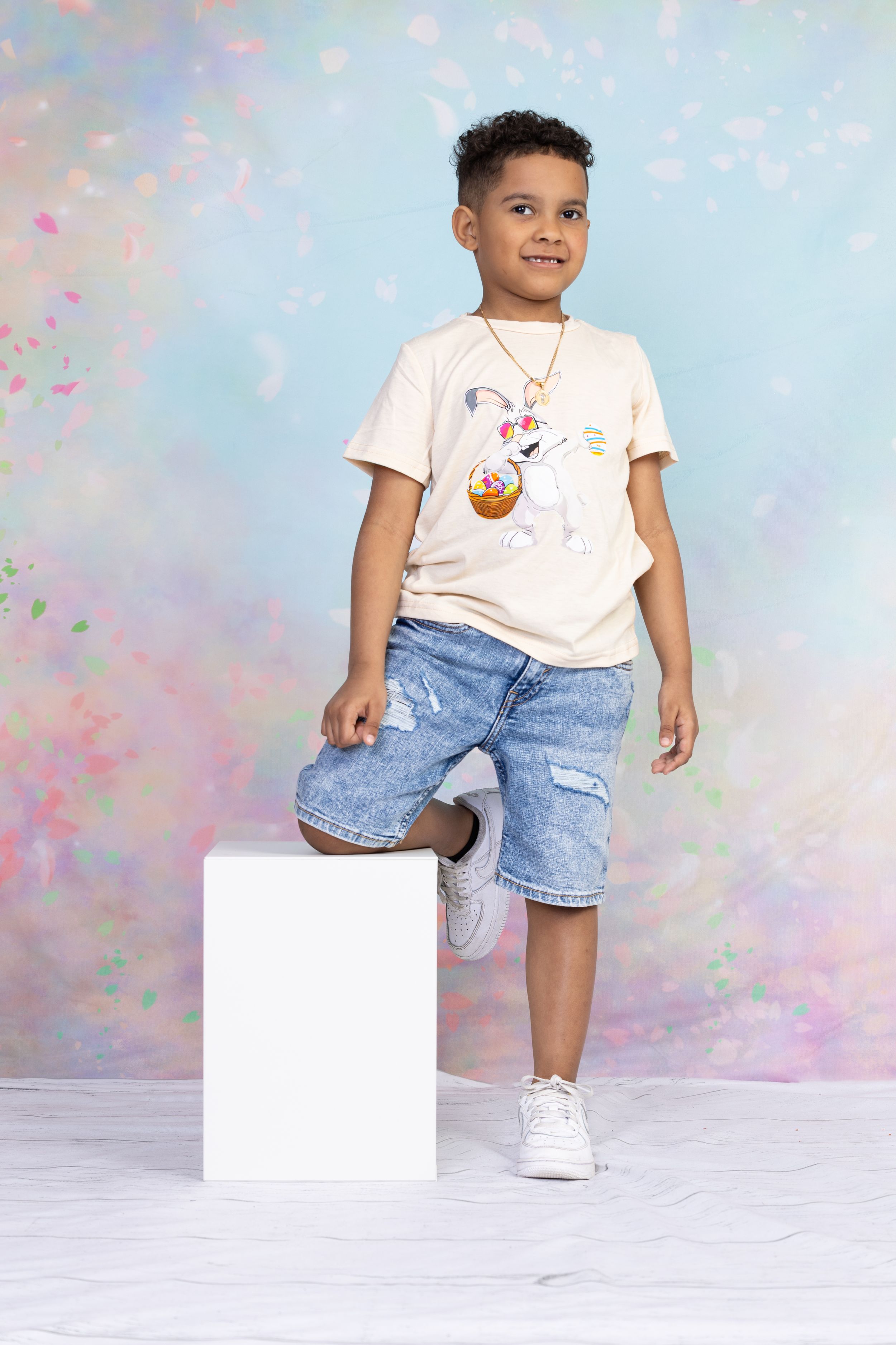 Young person in white bunny graphic tee and denim shorts posing with foot on white cube.