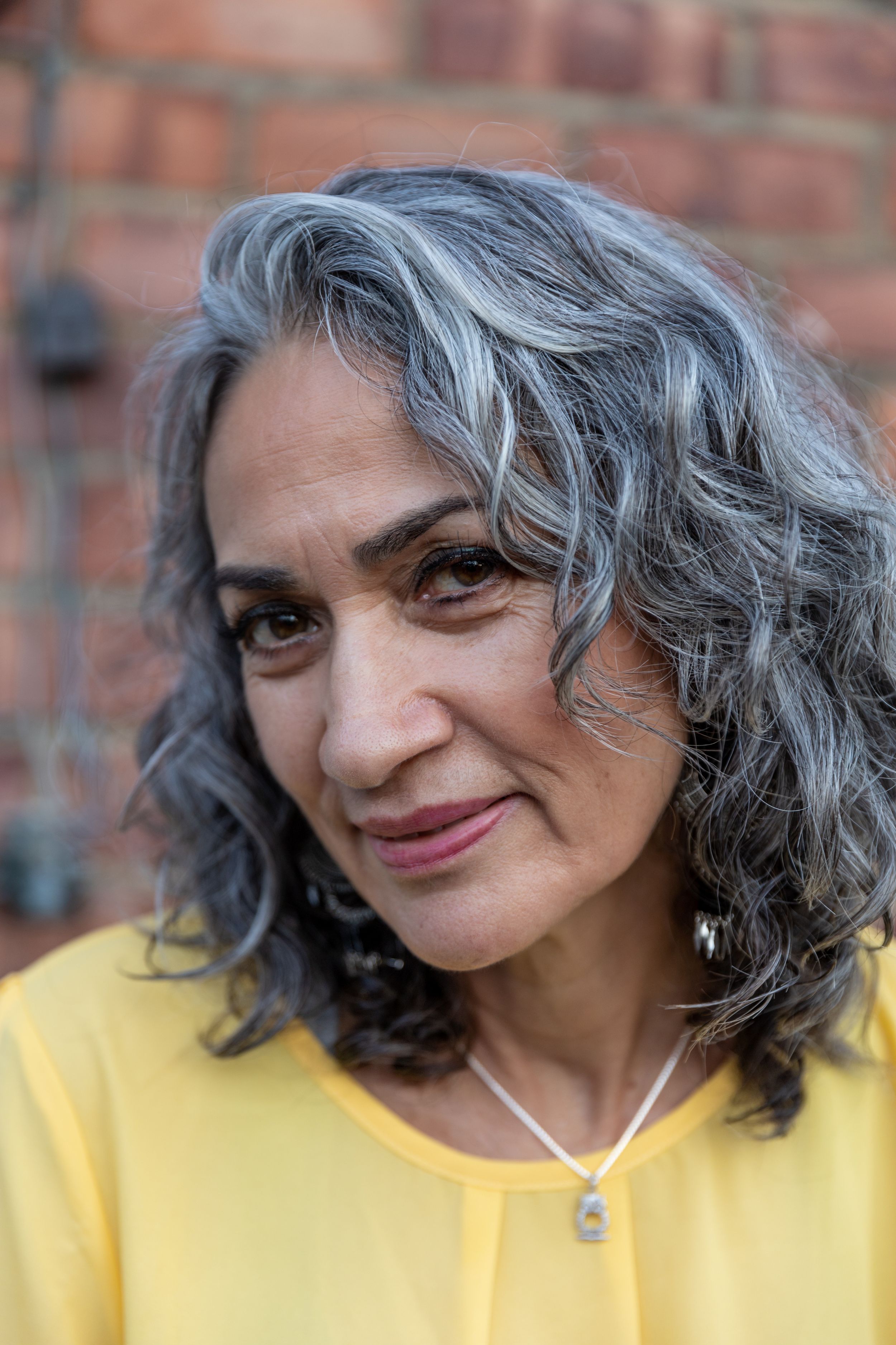 Portrait of person with silver curly hair wearing yellow top against brick background.