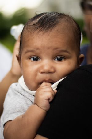 Close-up of baby in gray shirt with hand near mouth looking at camera.