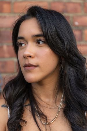 Portrait of person with long dark wavy hair wearing gold necklace against brick wall.