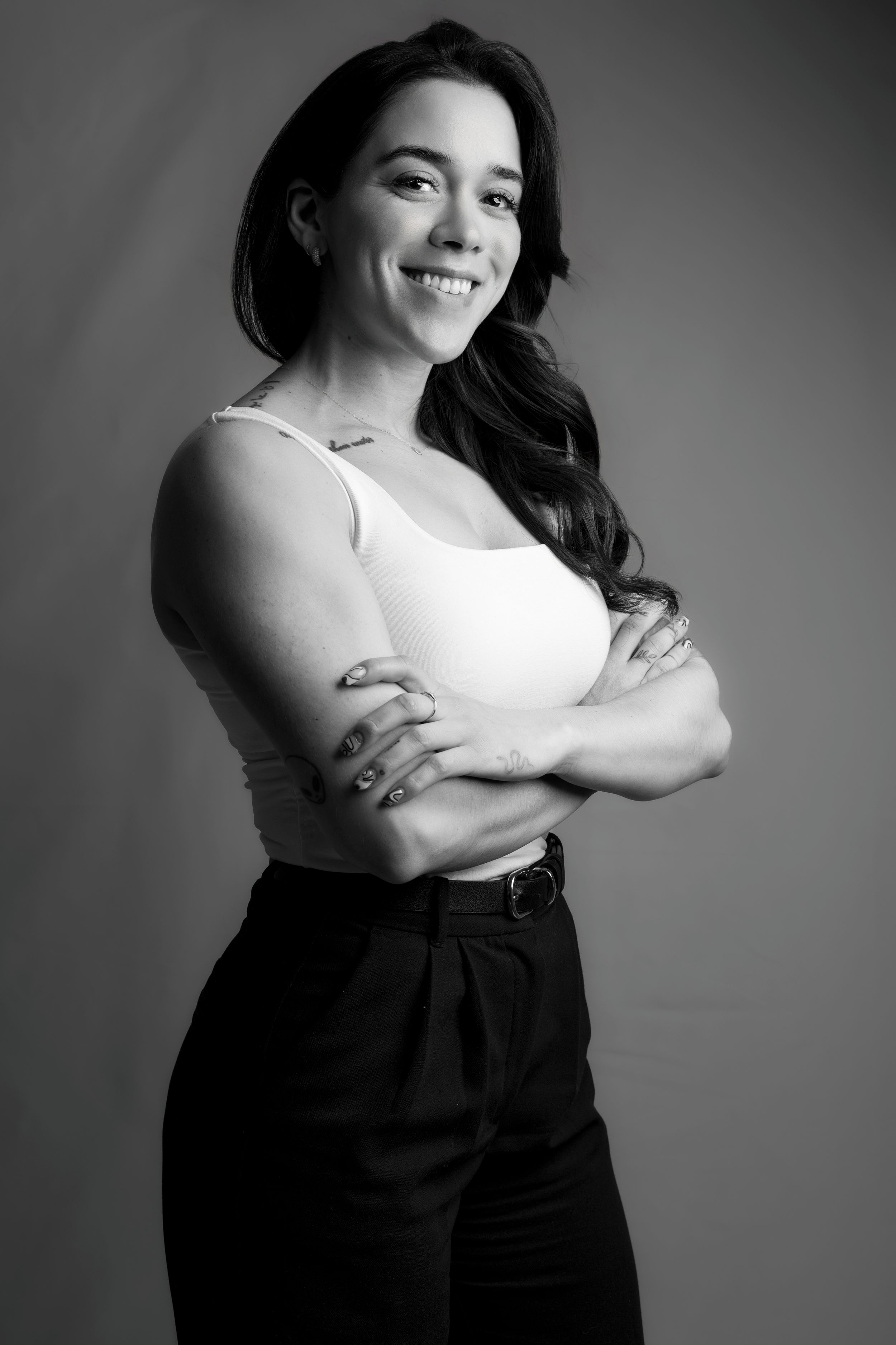 Black and white studio shot of woman in white top and dark pants showing arm tattoos.