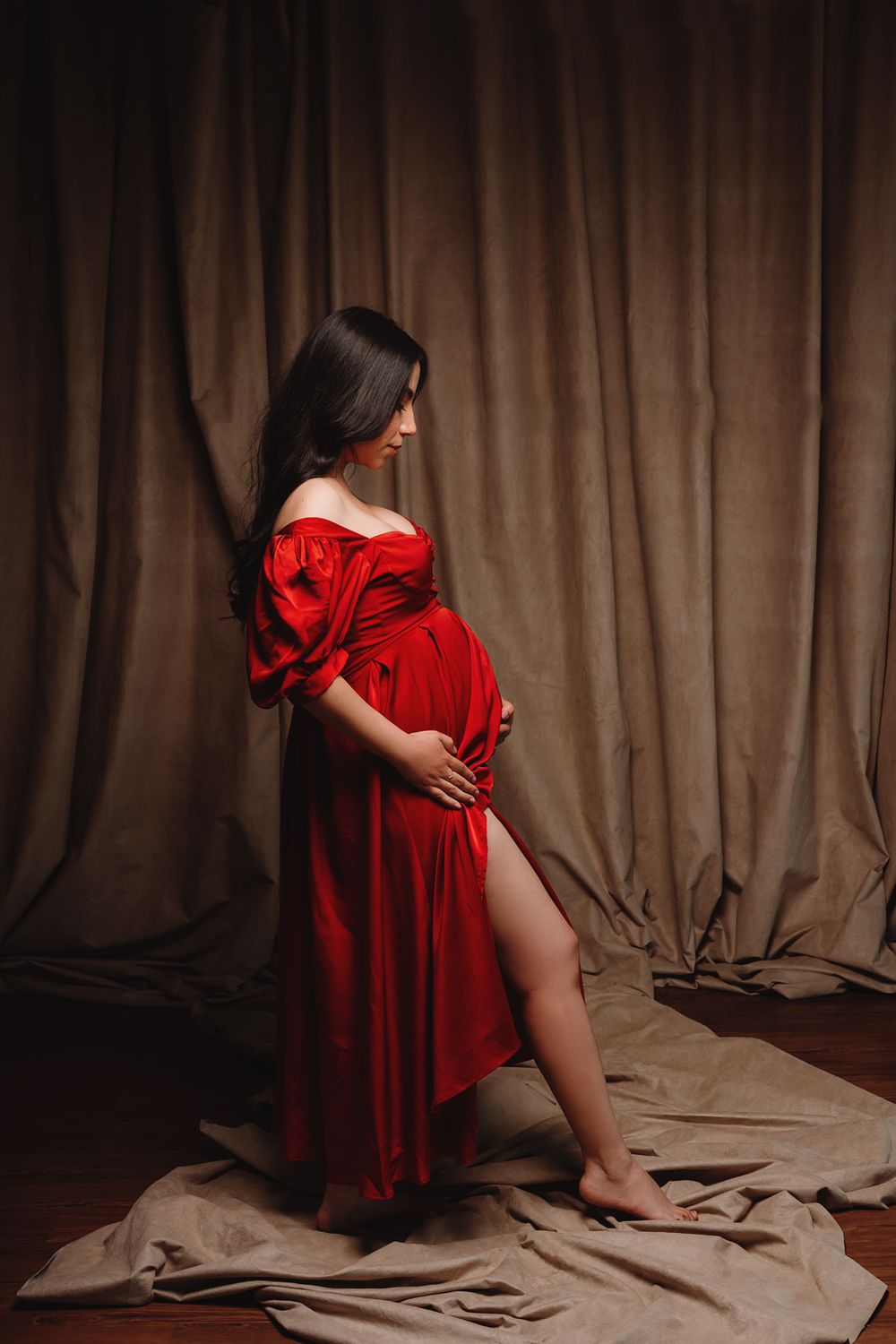 Expectant woman in flowing red velvet dress poses elegantly against dark curtain backdrop during maternity photoshoot.