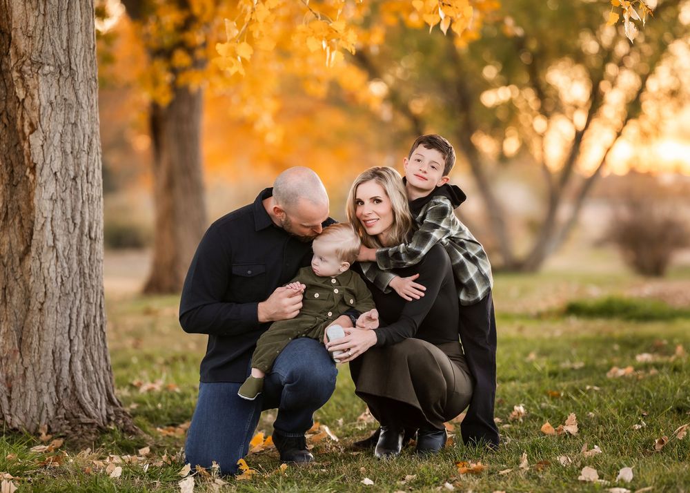 Family in dark clothing sits together on grass during golden autumn sunset with fallen leaves around them.
