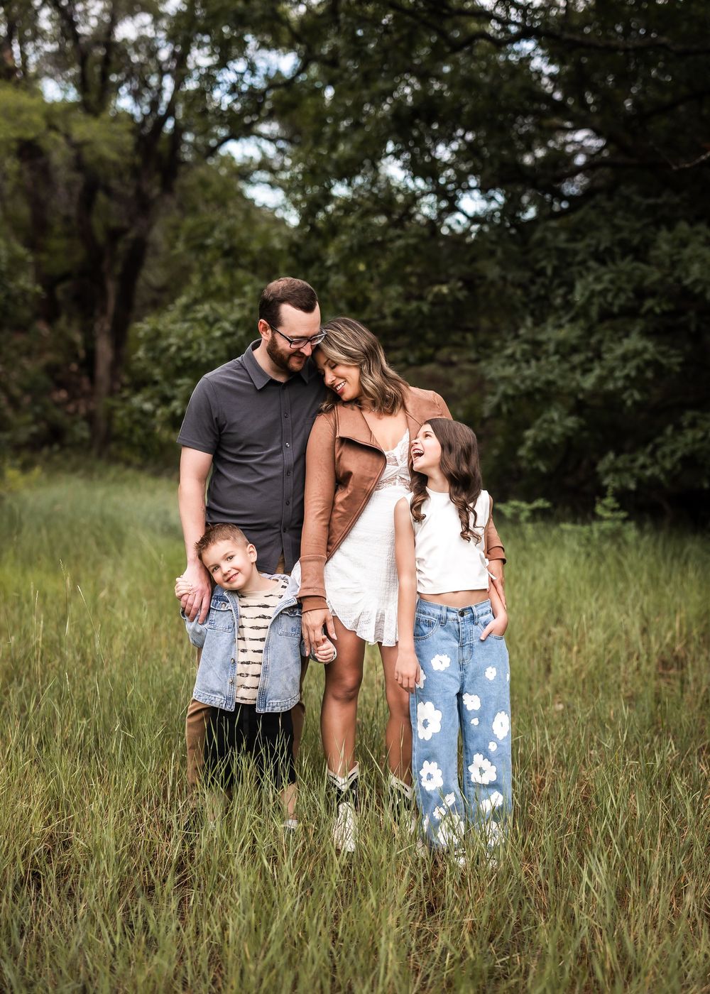 Family sharing candid moments together in a serene field surrounded by trees on a sunny day.
