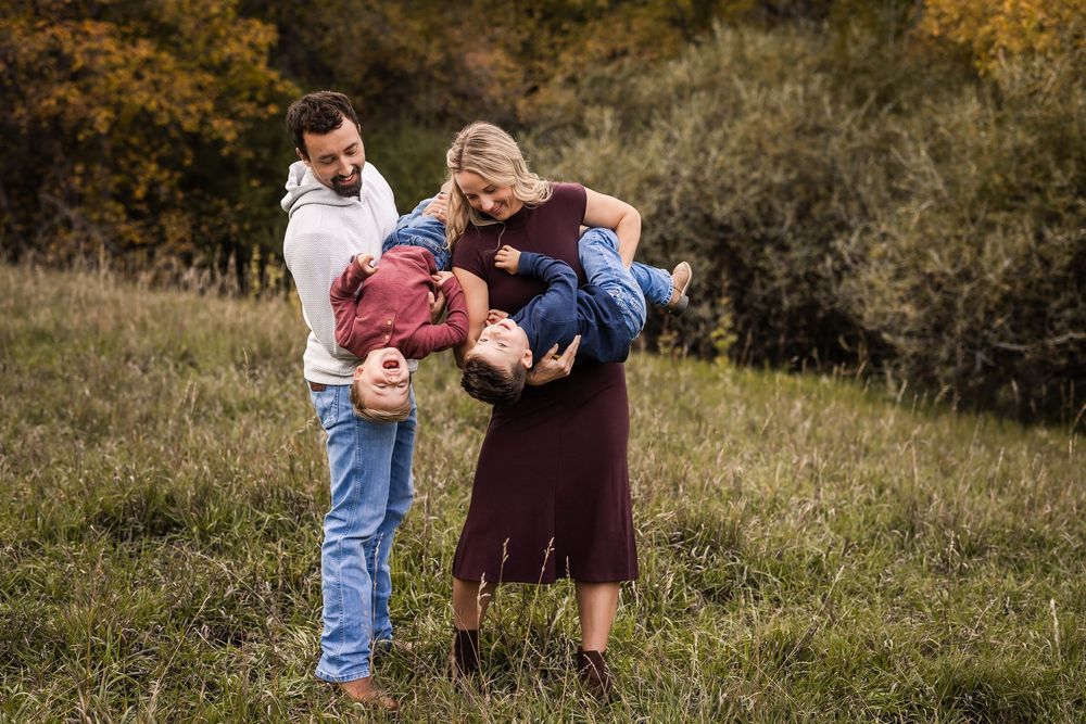 Family of four playing outdoors in autumn field, parents swinging two young children upside down while laughing together.