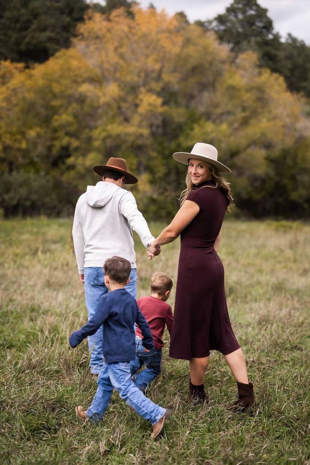 Family of four walking through autumn field, mother in burgundy dress and wide-brim hat looks back smiling, holding children's hands.