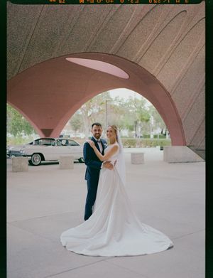 A wedding couple poses under a curved concrete archway at an outdoor venue with vintage cars in the background.