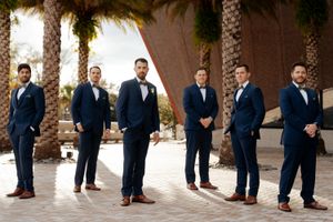 Groomsmen in navy suits and brown shoes pose in a line against stone columns at an outdoor venue.