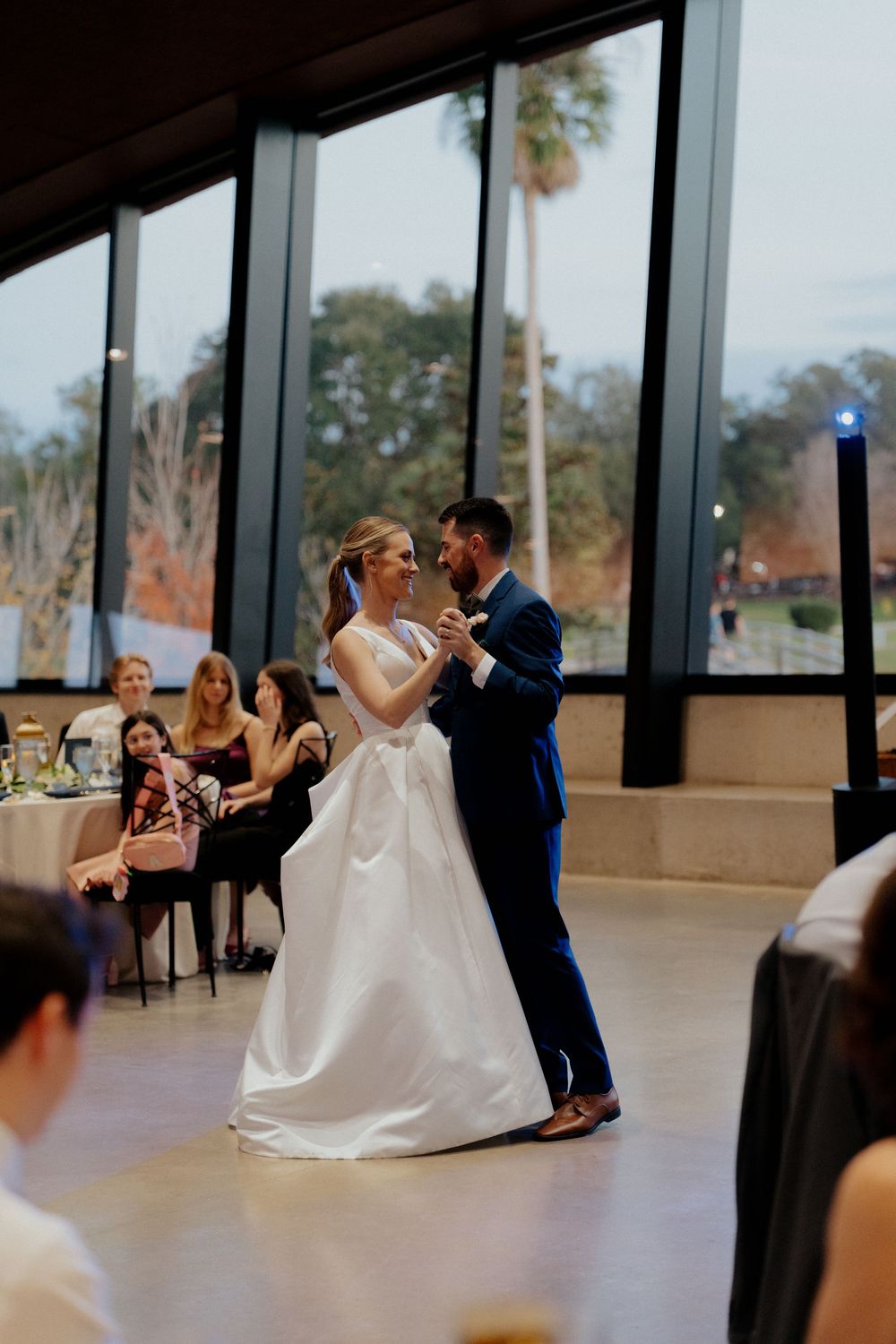 A couple dances together during their wedding reception in a bright venue with floor-to-ceiling windows.